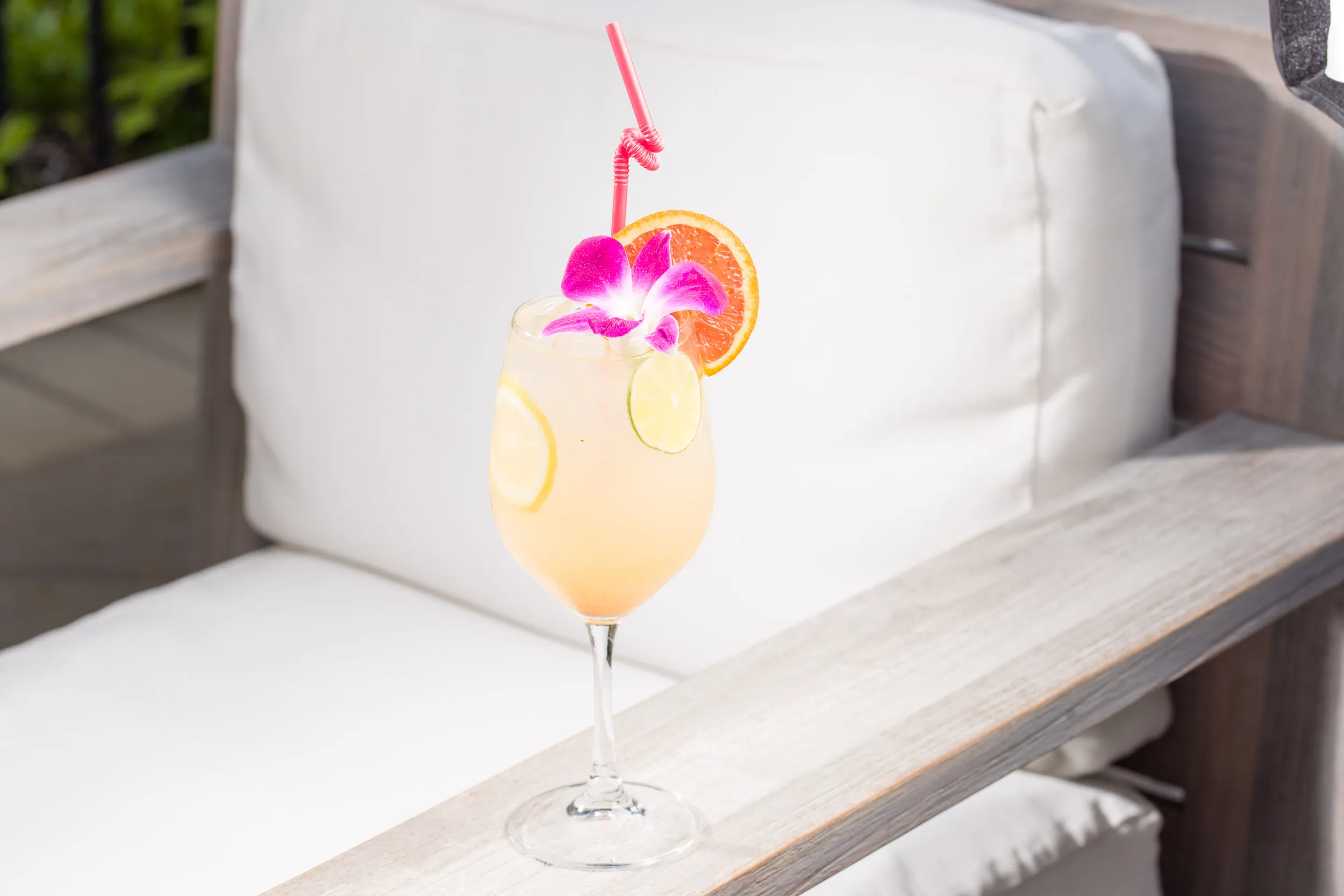 A glass of citrus cocktail with lemon slices, a pink straw, a purple orchid flower, and an orange slice garnish sits on the armrest of a white cushioned outdoor chair in bright sunlight.