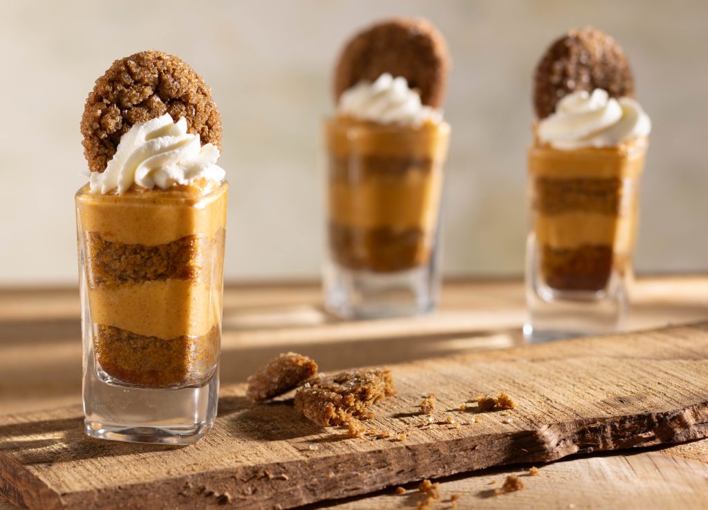 Three layered pumpkin parfaits in small glasses, topped with whipped cream and a gingersnap cookie, are arranged on a rustic wooden board with cookie crumbs scattered in the foreground.