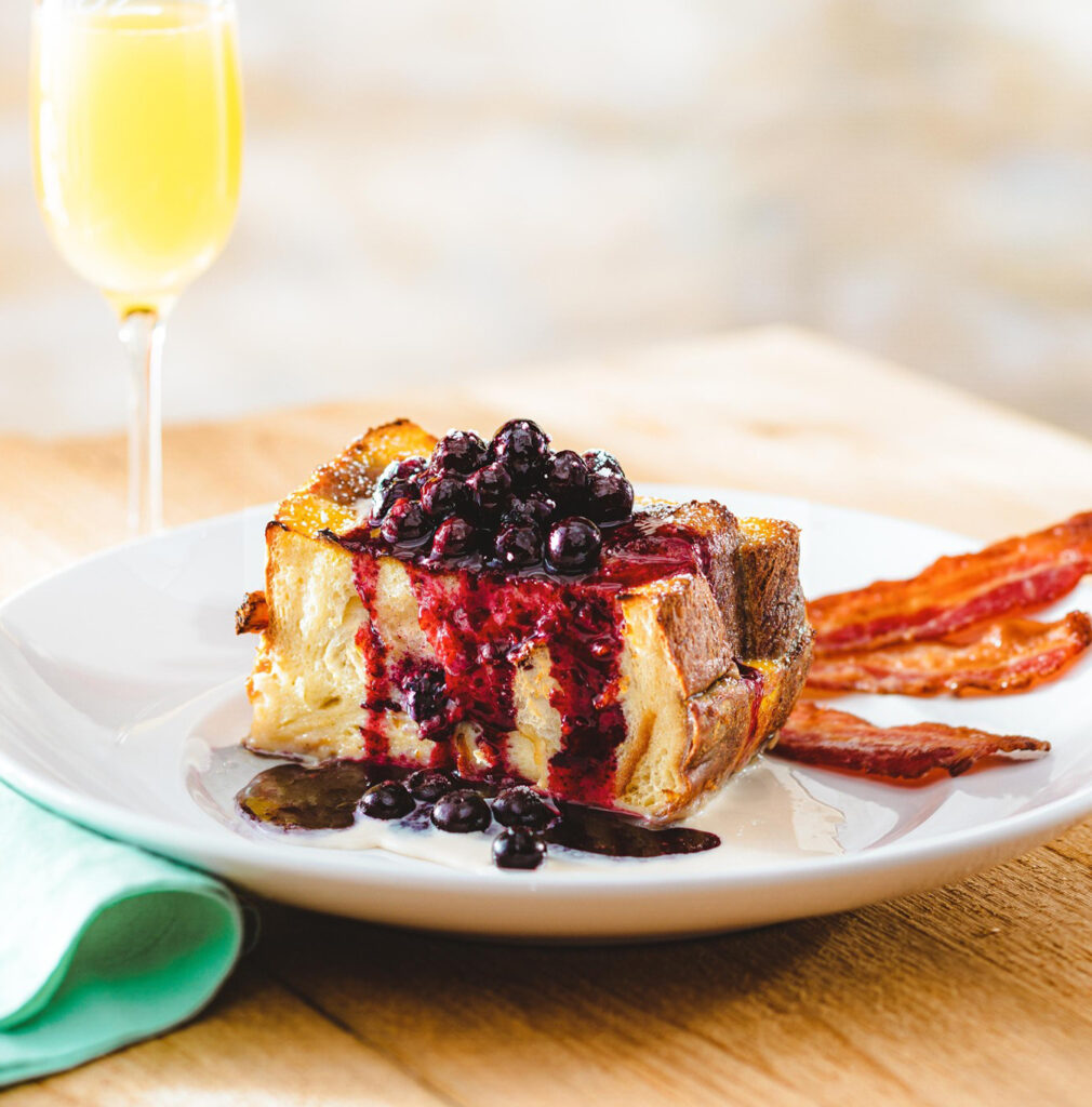 A thick slice of French toast topped with blueberries and sauce, served with strips of crispy bacon on a white plate. A glass of orange juice is in the background on a wooden table.