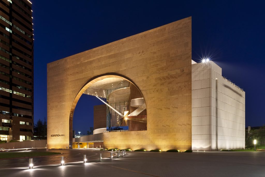 A modern building with a large, illuminated archway and geometric design, photographed at night. The structure has tan stone and white surfaces, with spotlights highlighting its architectural features.