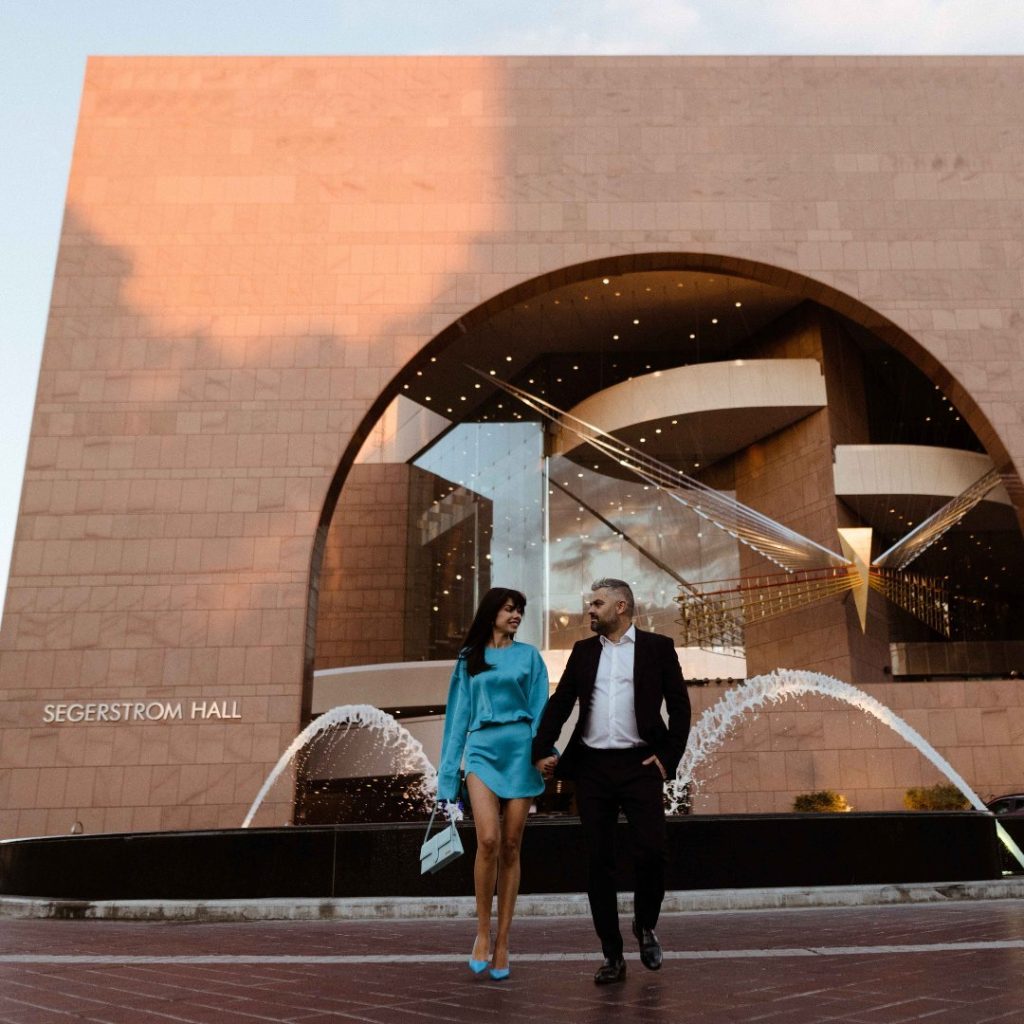 A well-dressed couple walks hand-in-hand in front of Segerstrom Hall, a modern building with a large arched entrance and water fountains spraying in the foreground.