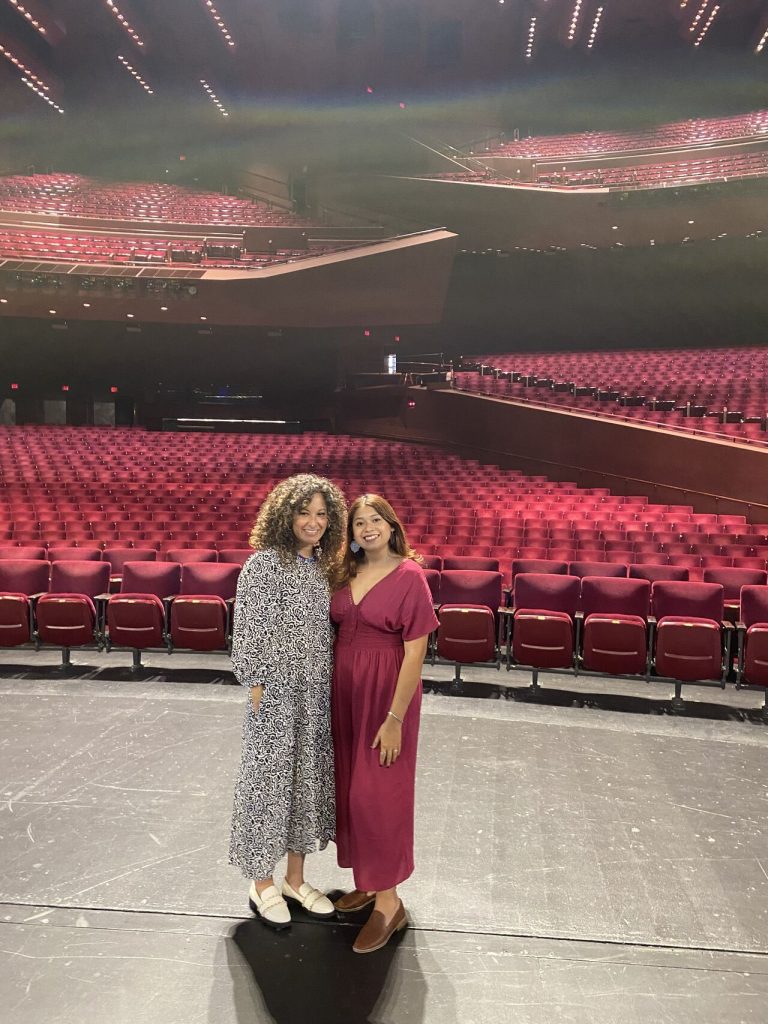 Two women stand together and smile on a theater stage, facing the camera. Rows of empty red seats and balconies fill the large auditorium behind them.