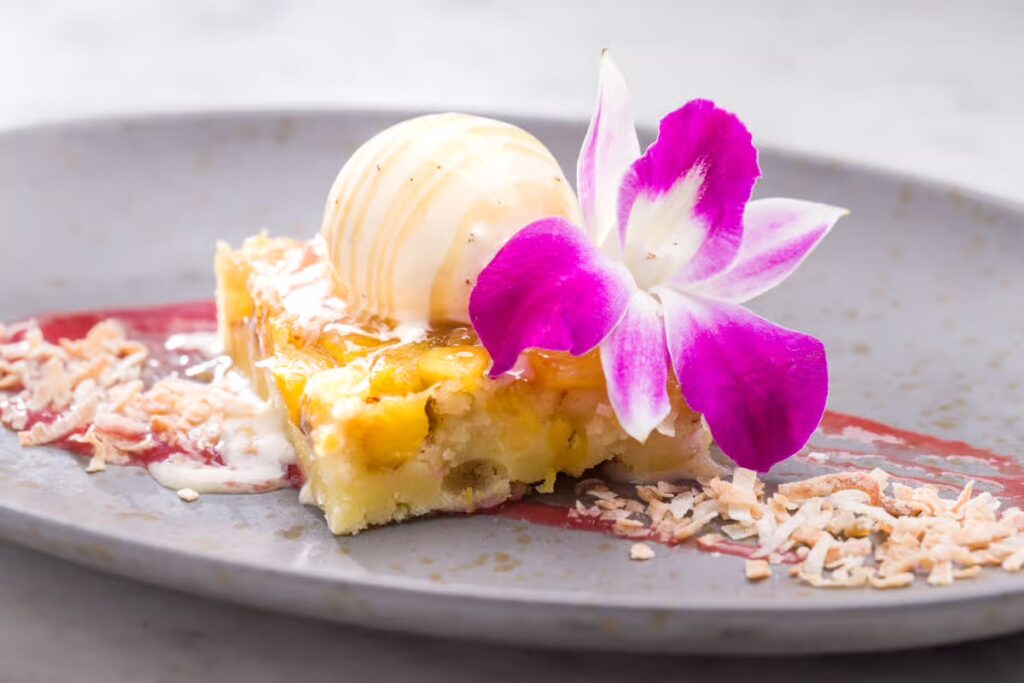 A plated dessert at Silver Trumpet Restaurant Costa Mesa features a square piece of cake, a scoop of vanilla ice cream, a purple and white orchid, toasted coconut flakes, and a drizzle of sauce on a gray plate.