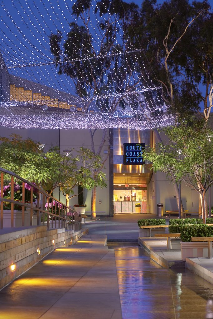 A well-lit walkway at night leads to the entrance of South Coast Plaza, with trees, decorative lights overhead, and a water feature alongside the path.