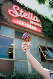 A person holds up a waffle cone with purple ice cream and rainbow sprinkles in front of a building with a red neon sign that reads Stella Jeans Ice Cream, reminiscent of sweet treats near Silver Trumpet Restaurant Costa Mesa.