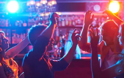 People dancing and raising their hands in a dimly lit nightclub or bar with colorful blue and red lights in the background. Bottles line the shelves behind the bar—a must-see spot featured on our Costa Mesa travel blog.