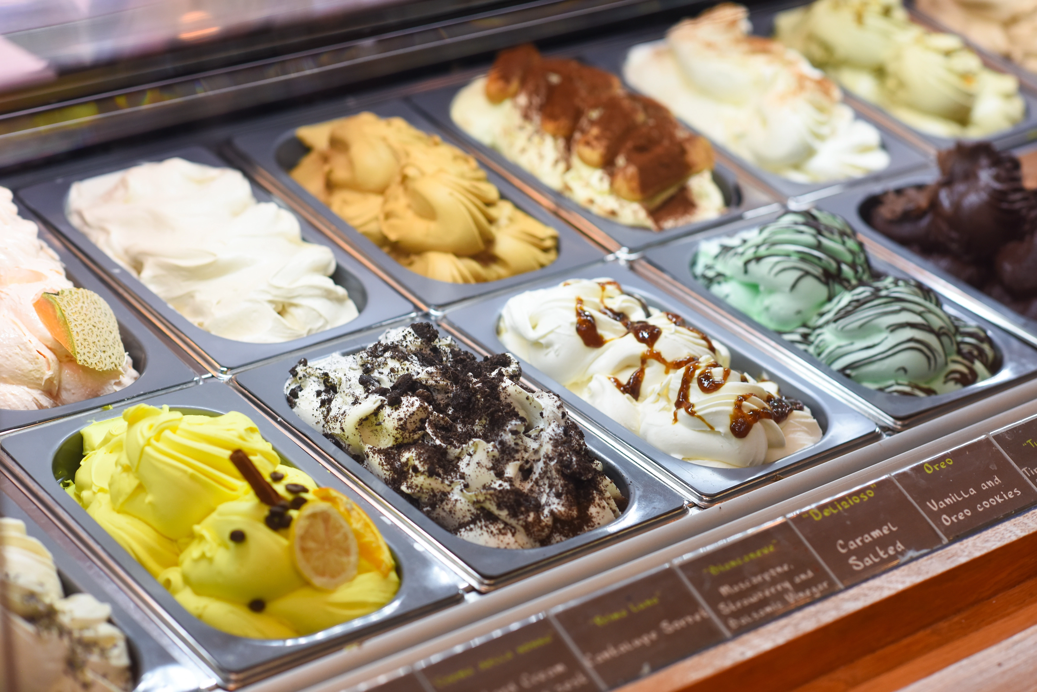 A display case filled with various flavors of gelato in metal trays, including lemon, mint, chocolate, and caramel—each decorated with matching toppings—a perfect highlight for any Costa Mesa travel blog.
