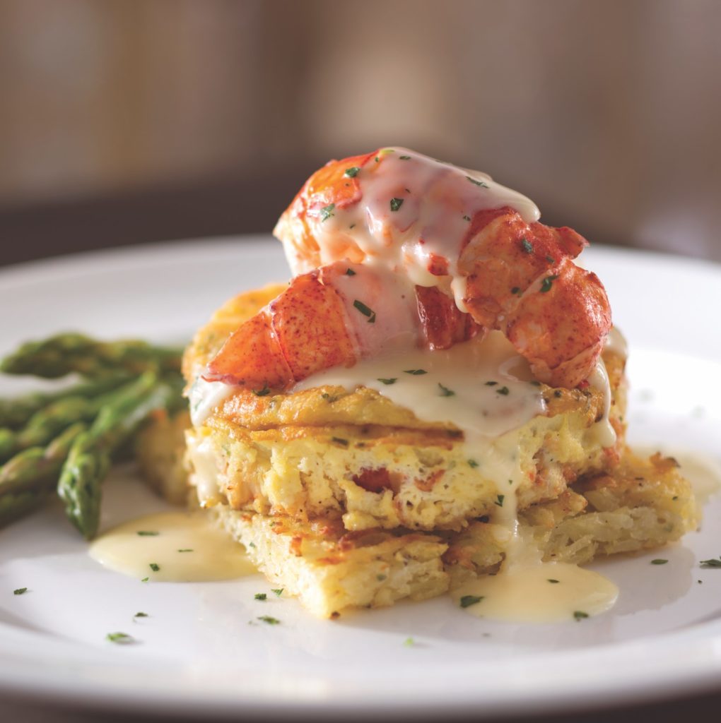 A gourmet dish featuring lobster claws on top of a creamy sauce and a savory potato cake, garnished with herbs, with asparagus spears on the side, served on a white plate.
