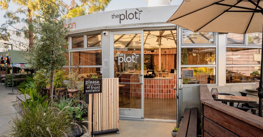 Exterior of the plot restaurant with glass doors, greenery, and a sign requesting guests to check in at the front entrance. Outdoor seating and potted plants are visible around the entryway.