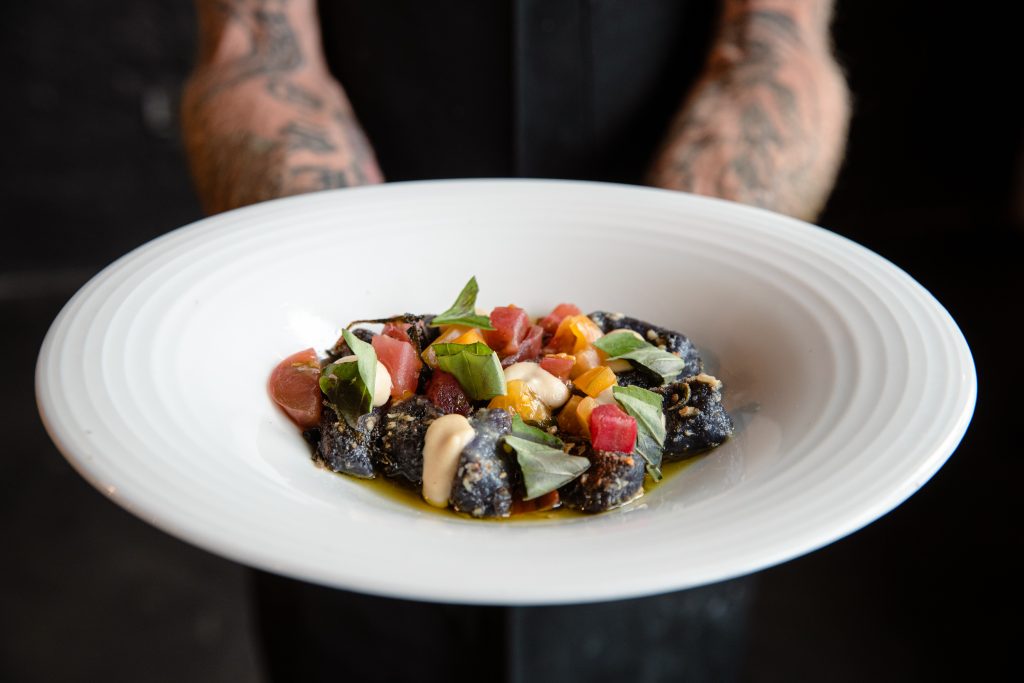 A tattooed person holds a white plate with a gourmet dish featuring dark-colored gnocchi, fresh herbs, colorful diced vegetables, and a creamy sauce, presented artfully.