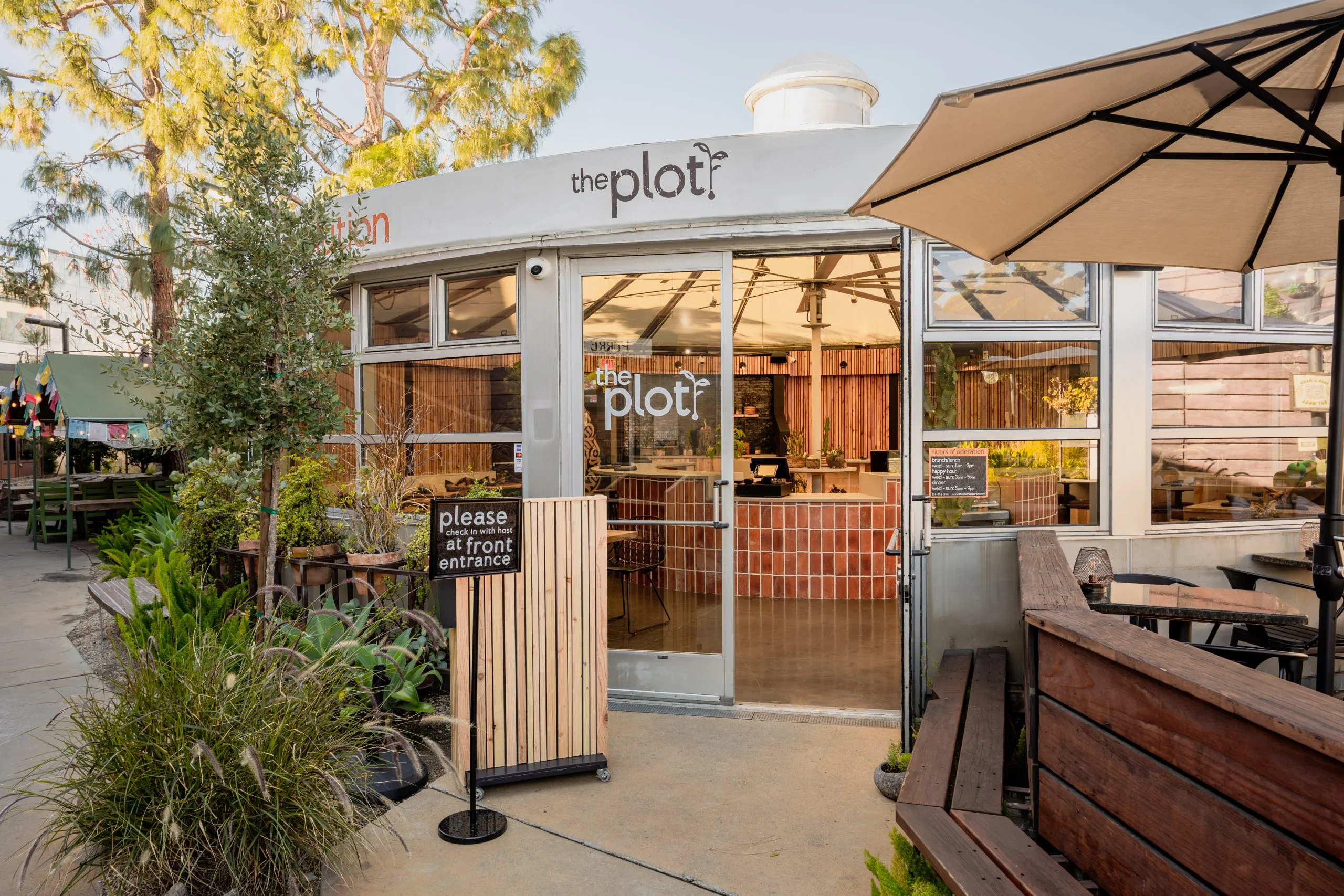 A modern restaurant entrance with glass doors labeled “the plot” welcomes visitors. Outdoor seating and plants create an inviting vibe, making it a perfect spot featured on any Costa Mesa travel blog. Sunlight brightens the well-marked front entrance.