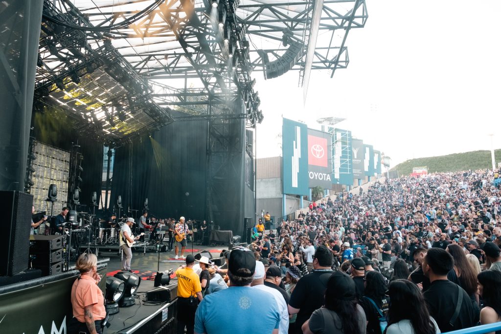 A large crowd watches a live band perform on an outdoor stage at an amphitheater during daylight. The audience stands close to the stage, and big screens and sponsor signs are visible in the background.