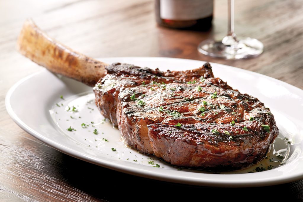 A grilled tomahawk steak garnished with chopped herbs sits on a white plate, with a wine glass and bottle blurred in the background on a wooden table.