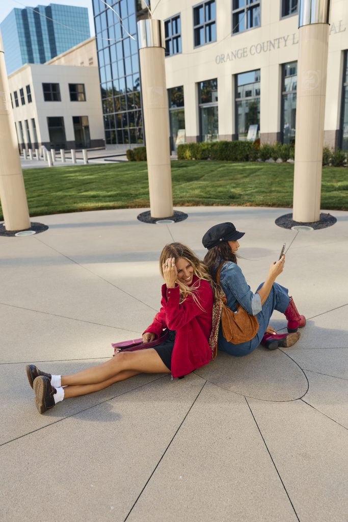 Two women sit back-to-back on a paved area outside the Orange County Legal building. One woman smiles at the camera, while the other looks at her phone. Both wear casual outfits and appear relaxed.