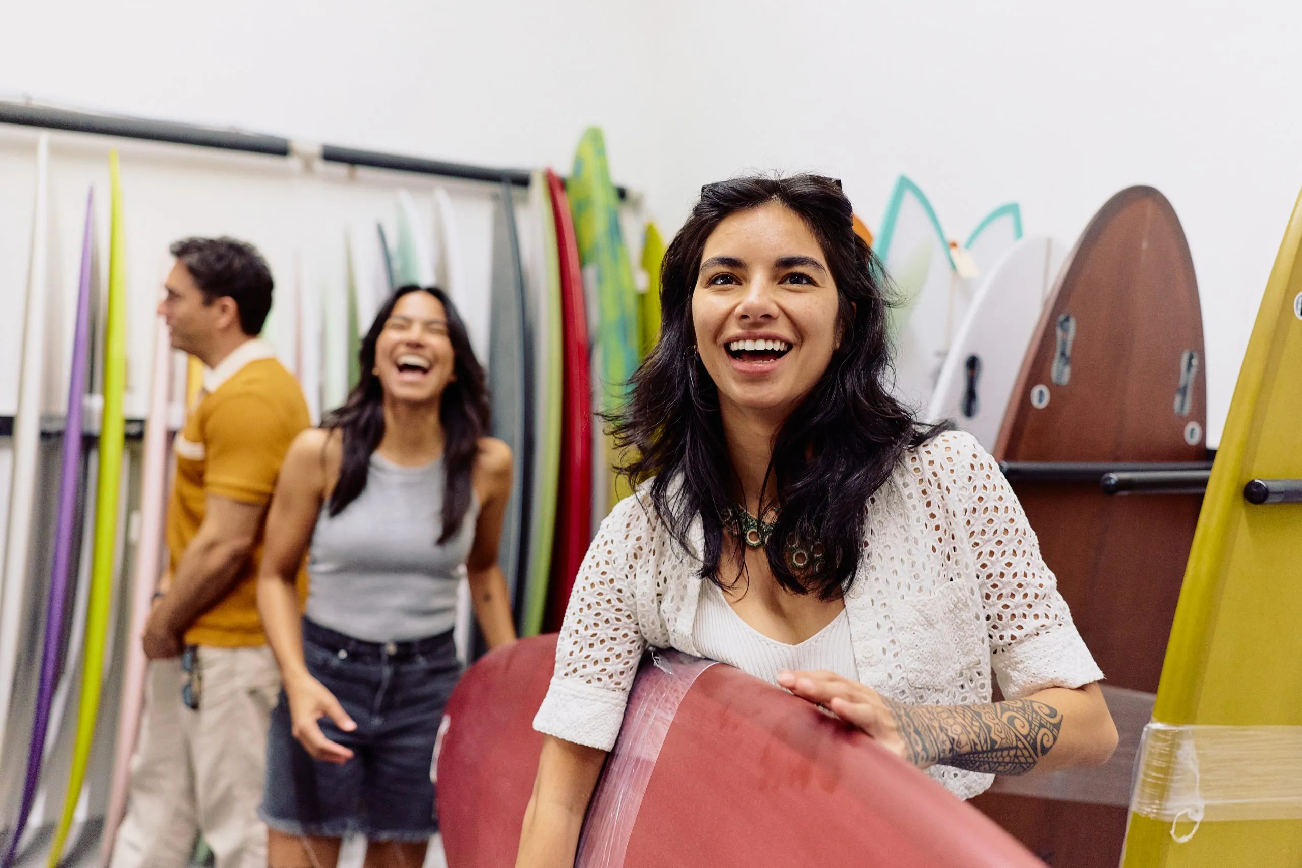 Three people smile and laugh in a surf shop, surrounded by colorful surfboards. Perfect for a Costa Mesa travel blog, a woman in front holds a red surfboard while her friends stand behind her, all appearing cheerful and relaxed.