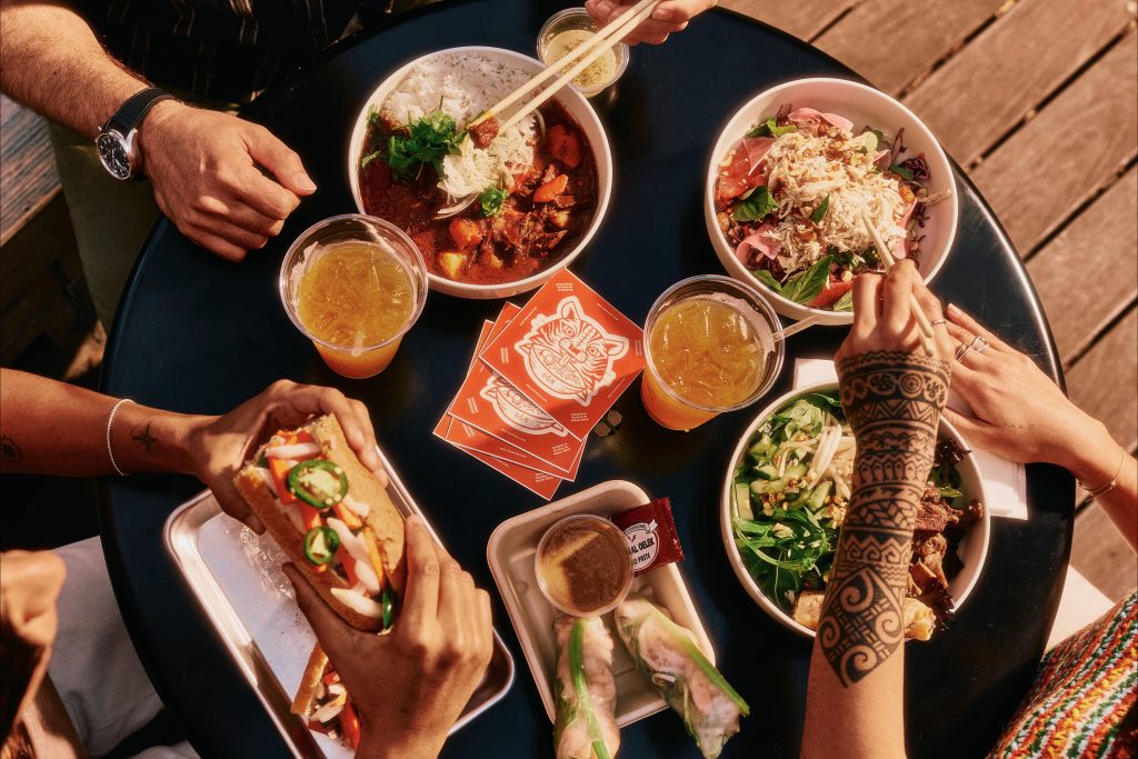 A top view of a table with bowls of food, drinks, spring rolls, and a sandwich. Several hands are reaching for the food, one with a henna tattoo, and three orange cards with a white fox design are on the table.