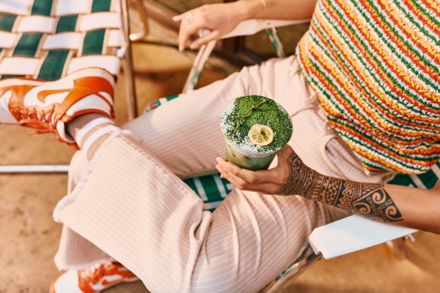 A person with a tattooed arm sits on a striped lawn chair, wearing peach pants, a colorful striped top, and orange sneakers, holding a green matcha drink topped with a banana slice and whipped cream.