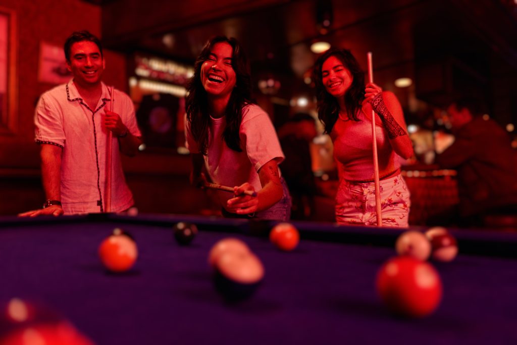 Three young adults smiling and playing pool in a warmly lit bar, with a focus on the billiard balls and cue sticks as they enjoy their game together.