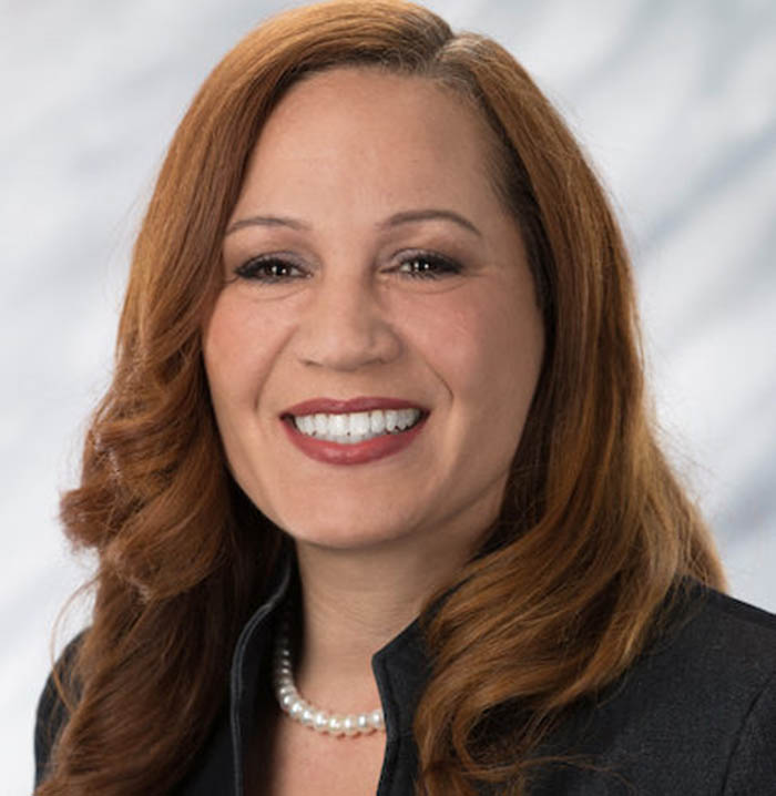 A woman with long, wavy auburn hair is smiling. She is wearing a dark blazer, a white pearl necklace, and has a light background behind her.