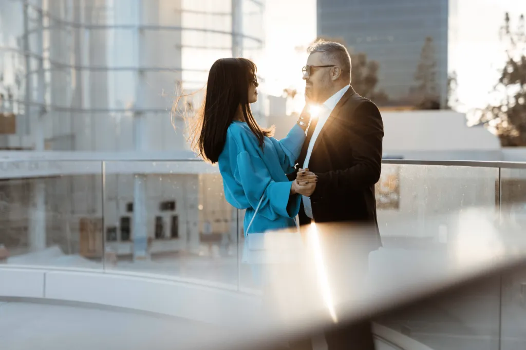 A man and woman stand close together on an outdoor balcony at sunset, smiling and holding hands. The sun shines brightly behind them, creating a warm and romantic atmosphere.