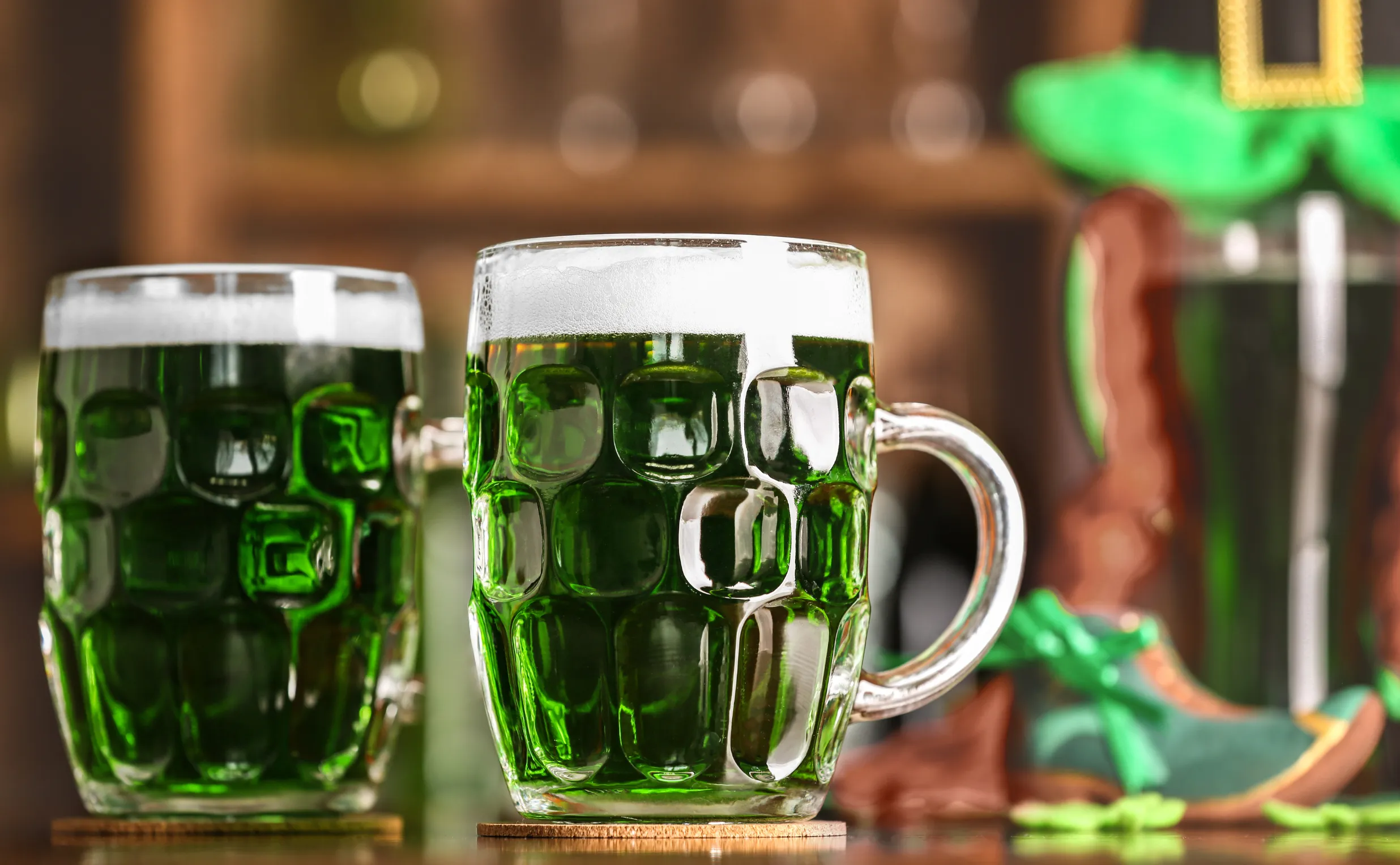 Two glass mugs filled with green beer sit on a bar, suggesting a festive St. Patrick's Day atmosphere—a scene perfect for sharing on a Costa Mesa travel blog. The background features blurred green and brown decorations, adding to the celebration.