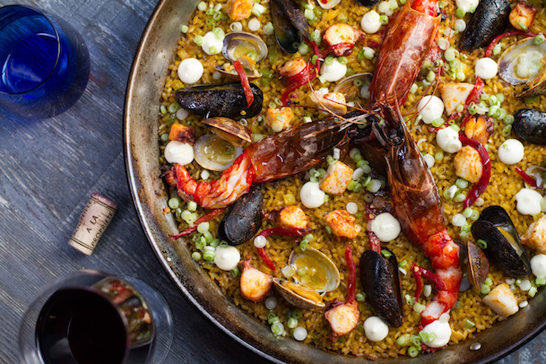 A close-up of seafood paella at Vaca Restaurant Costa Mesa, topped with shrimp, mussels, lobster, white sauce, and green peas. A glass of red wine, a blue glass, and a cork sit on the wooden table nearby.