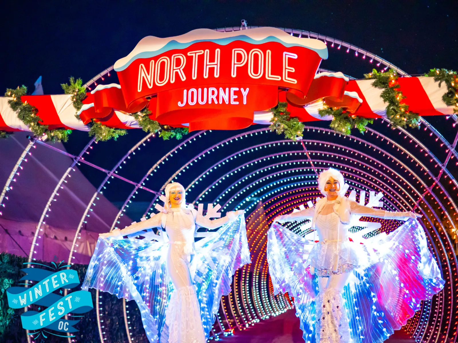 Two performers in illuminated white costumes pose under a festive North Pole Journey sign with candy cane decorations at night, perfect for a Costa Mesa travel blog. A Winter Fest OC logo is visible in the lower left corner.