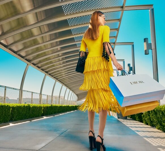 A woman in a bright yellow, tiered dress and black heels walks outdoors under a modern, curved canopy in Costa Mesa, carrying DIOR shopping bags and a black handbag on a sunny day.