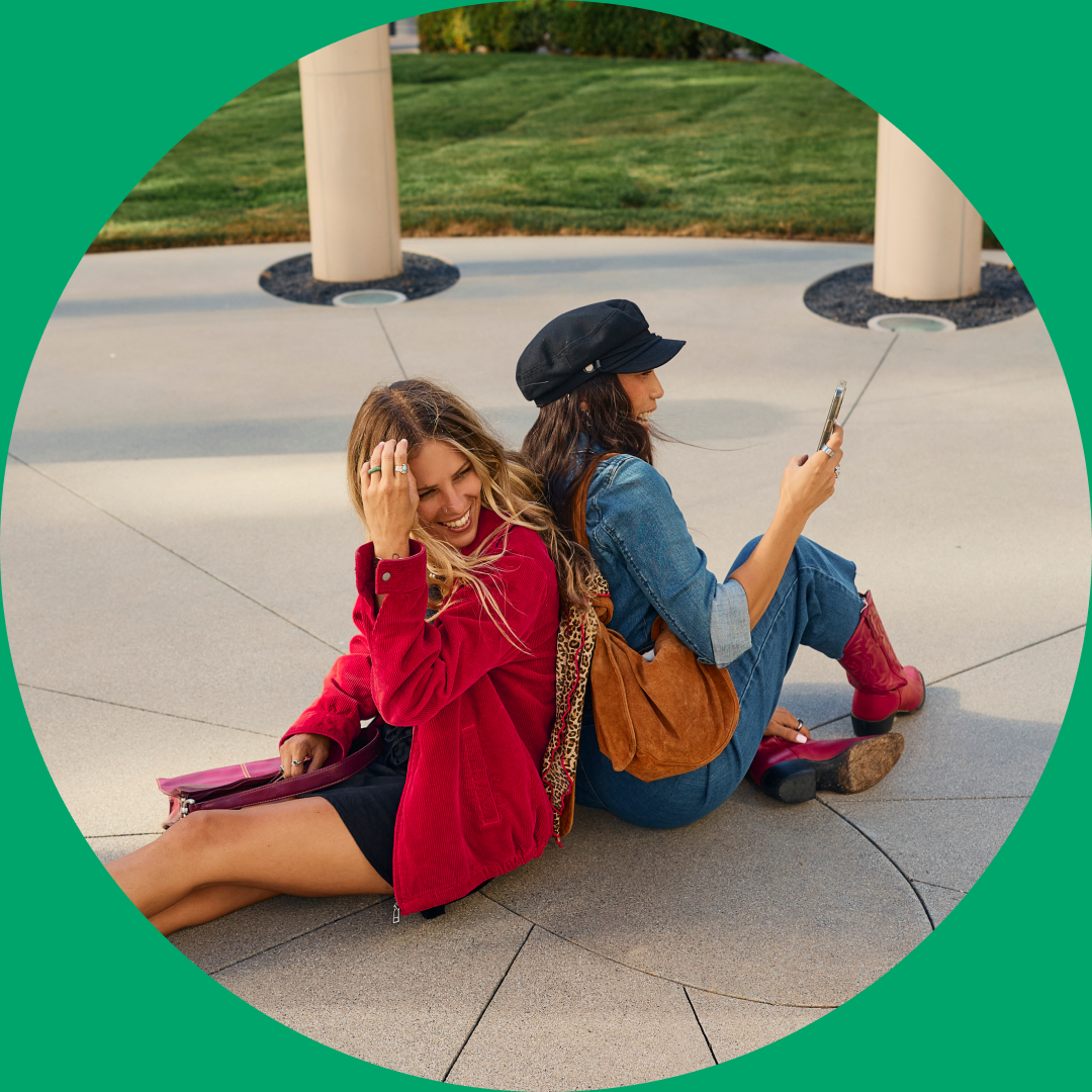 Two women sit back-to-back on a paved surface outdoors in Costa Mesa. One, in a red jacket, smiles at the camera, while the other, in denim and a black hat, checks her phone. Both have bags and are relaxed. A green border circles the image.