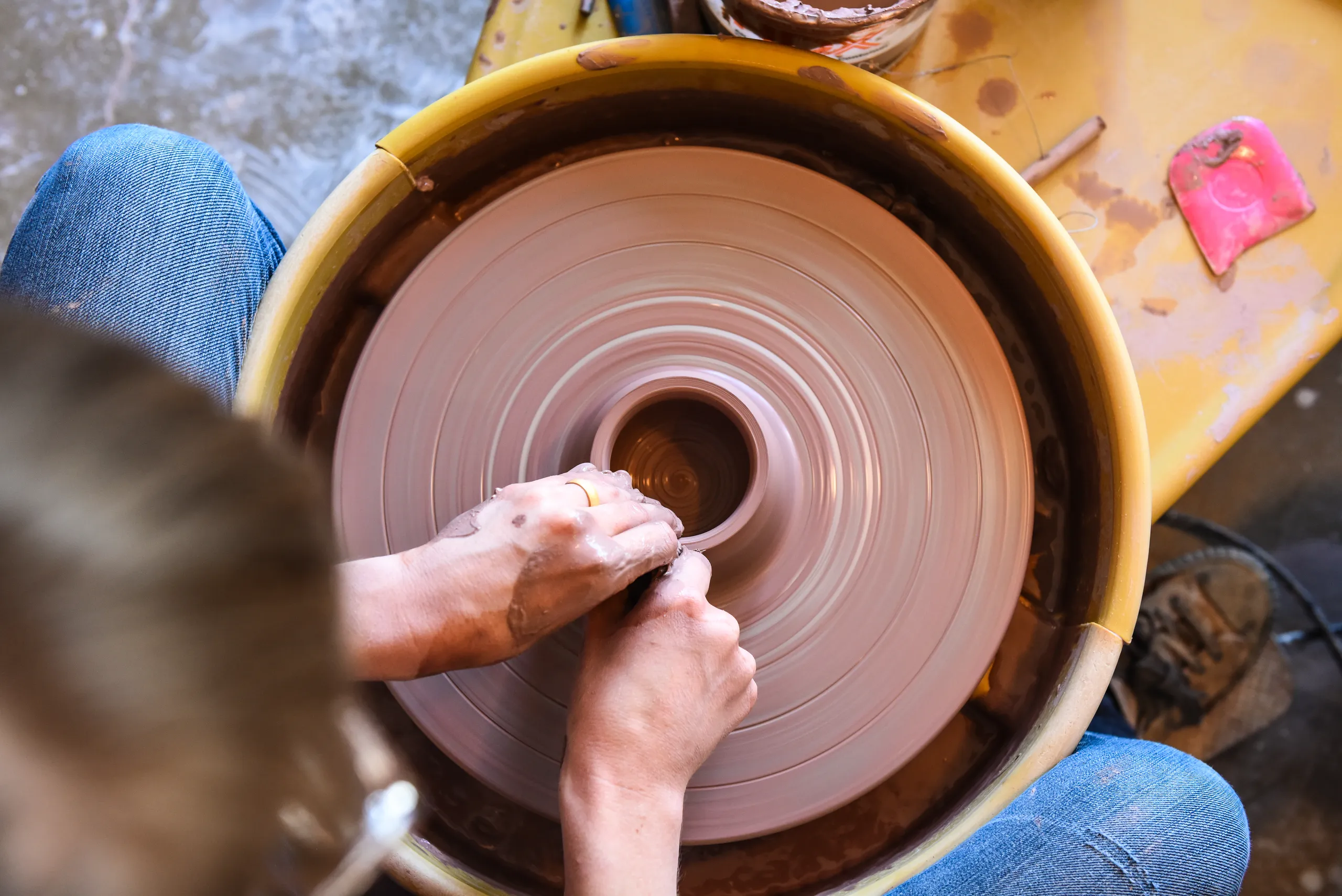 Person shaping clay on a pottery wheel with both hands, viewed from above. The spinning wheel and wet clay are the focus, with the persons arms and jeans partially visible.