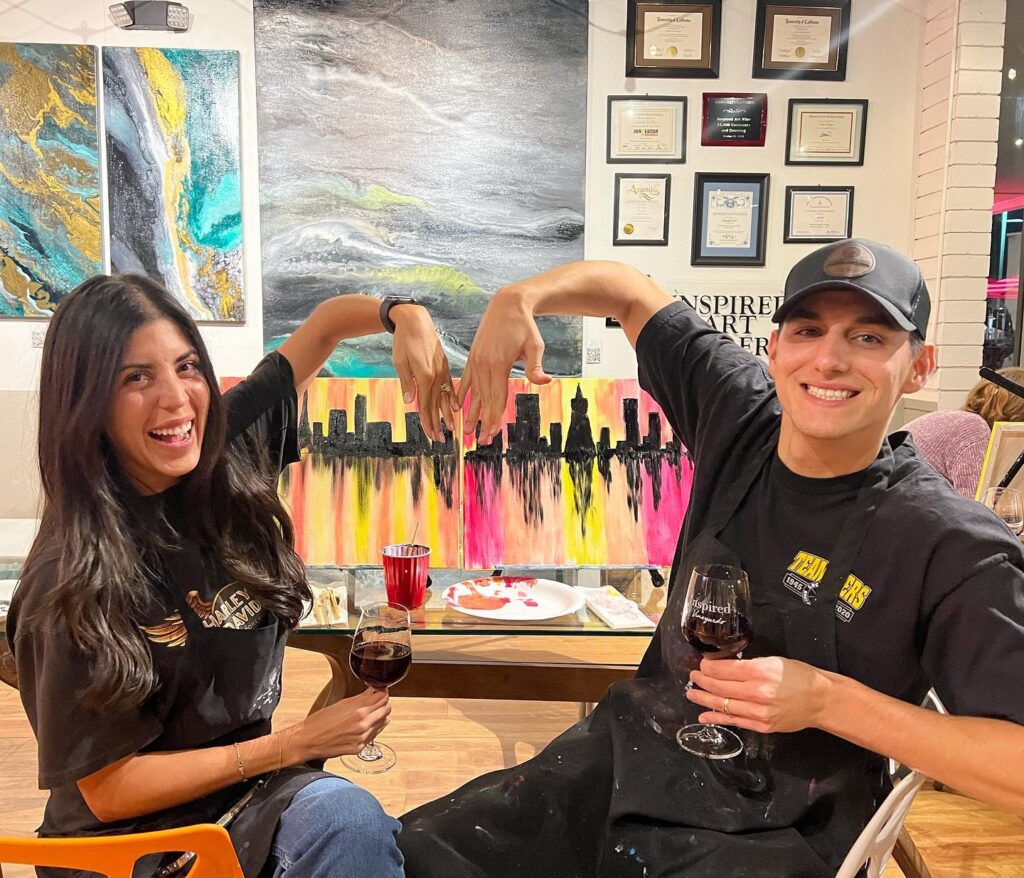 Two people smile and hold glasses of red wine while forming a heart shape with their arms in front of a colorful skyline painting at an art studio. Artworks and award certificates are displayed on the wall behind them.
