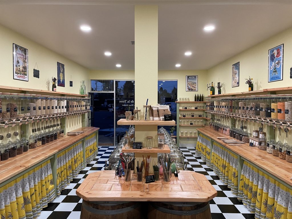 A well-lit zero-waste store with wooden shelves and bins filled with bulk food items, glass jars, and dispensers. Vintage posters decorate the yellow walls, and the floor has a black and white checkered pattern.