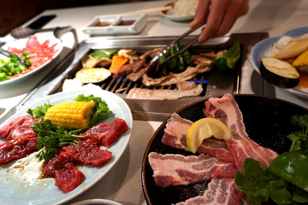 A person grills meat and vegetables on a tabletop barbecue, surrounded by plates of raw meat, corn, and greens, with dipping sauces and rice in the background.