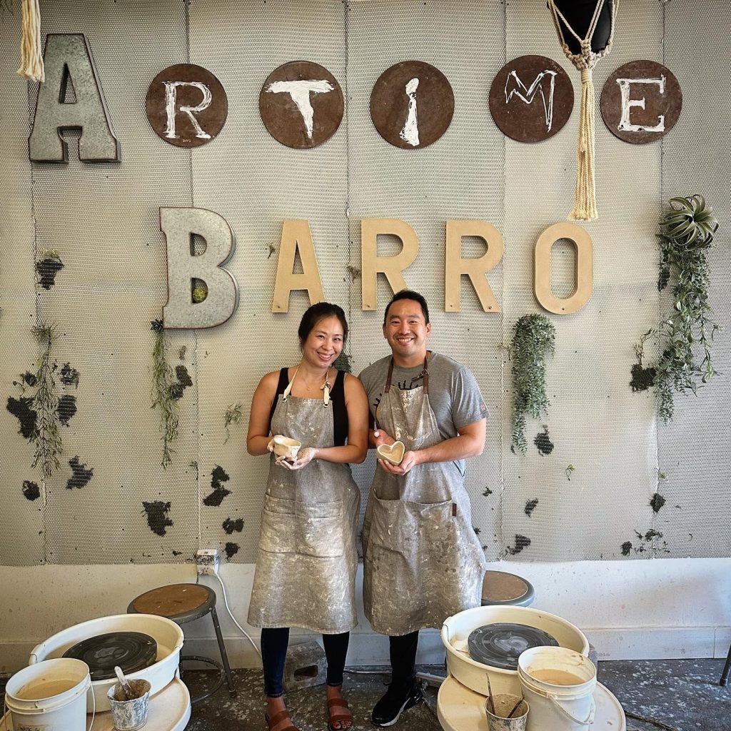 Two people wearing aprons stand and smile in a pottery studio, holding clay pieces. Behind them, large letters on the wall spell ARTIME BARRO. Pottery wheels and stools are visible in the foreground.