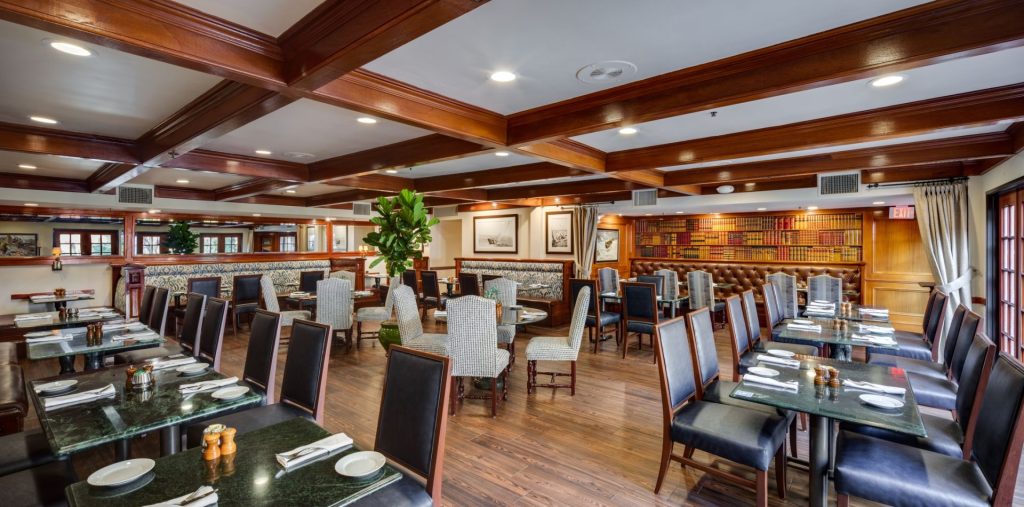 Spacious restaurant dining room with dark wooden floors, neatly set tables, cushioned chairs, wall art, large windows, and a bookcase theme on one wall under warm ceiling lighting.
