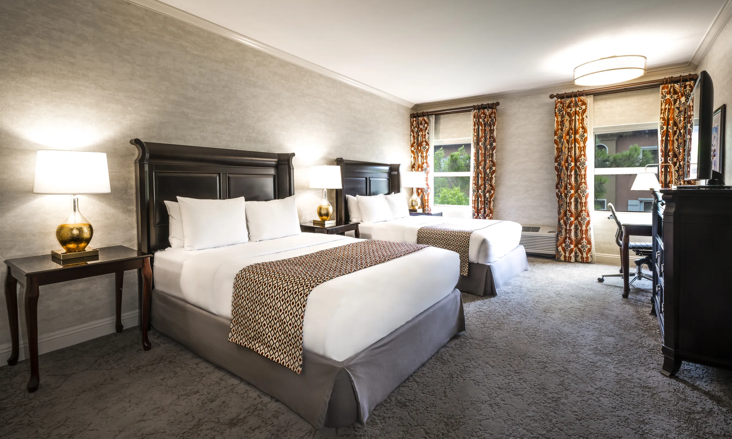Experience comfort and style in this Ayres Hotels room, featuring two queen beds with white bedding and patterned runners, nightstands with lamps, a desk with a chair, a dresser, and windows adorned with patterned curtains that allow plenty of natural light to brighten the space.