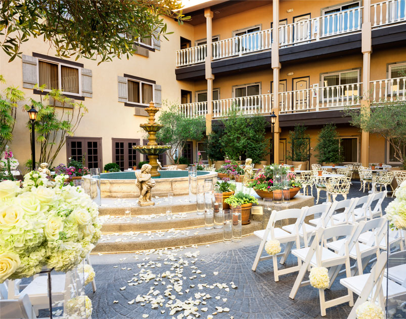 An elegant outdoor courtyard at Ayres Hotels set up for a wedding, featuring a central tiered fountain, white chairs with floral decorations, and flower petals along the aisle, all surrounded by lush greenery and a multi-story building.