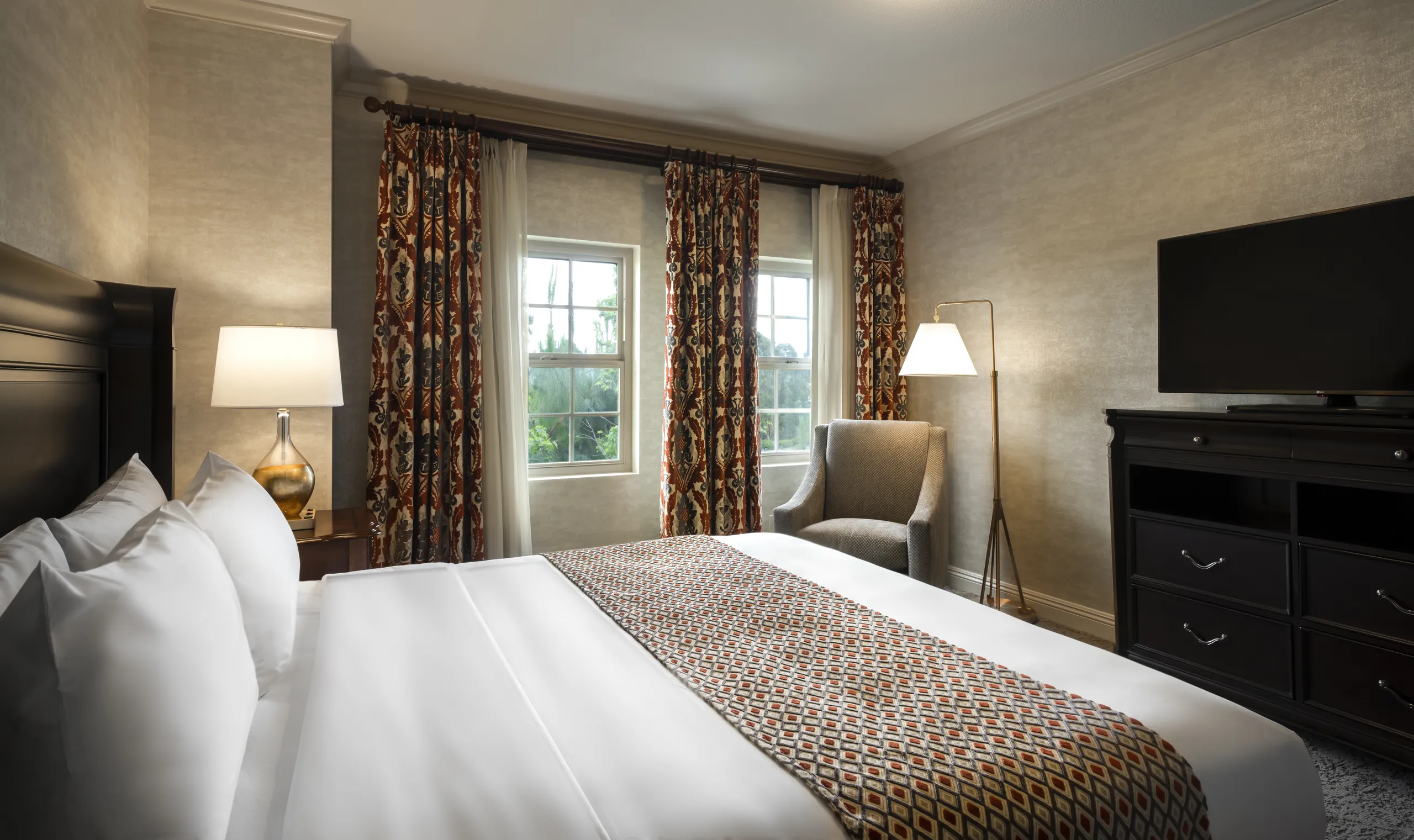 A neatly made Ayres Hotels room with a king bed, patterned throw, armchair, floor lamp, dresser with TV, and two windows with patterned curtains letting in daylight.