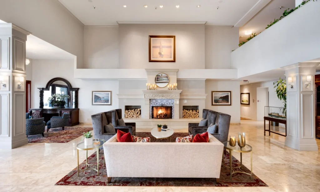 Elegant Ayres Hotels lobby with a fireplace, gray armchairs, a light sofa with red pillows, and coffee tables. The room features marble floors, large columns, artwork, and a cozy seating area with warm lighting.
