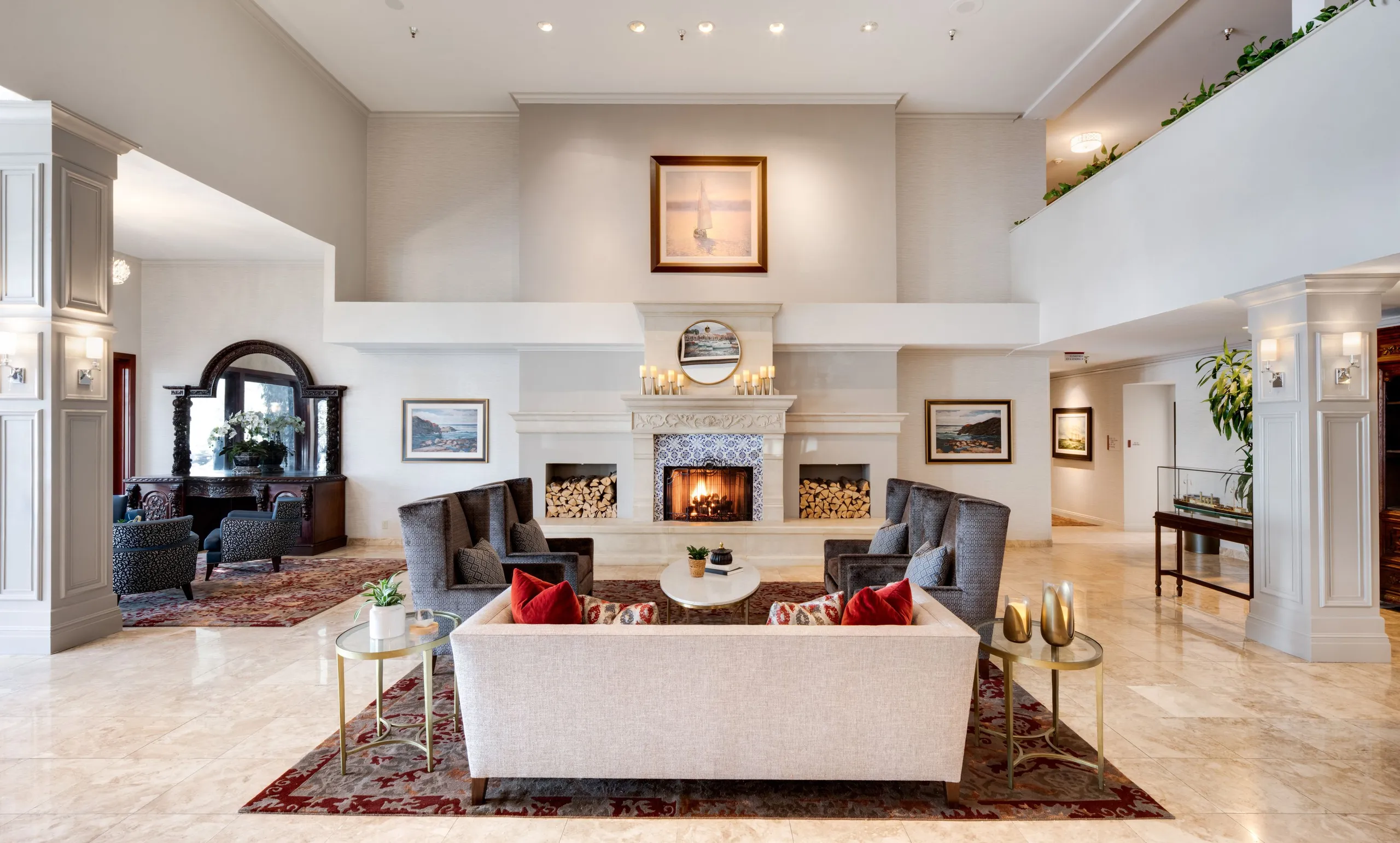 Elegant Ayres Hotels lobby with a fireplace, gray armchairs, a light sofa with red pillows, and coffee tables. The room features marble floors, large columns, artwork, and a cozy seating area with warm lighting.