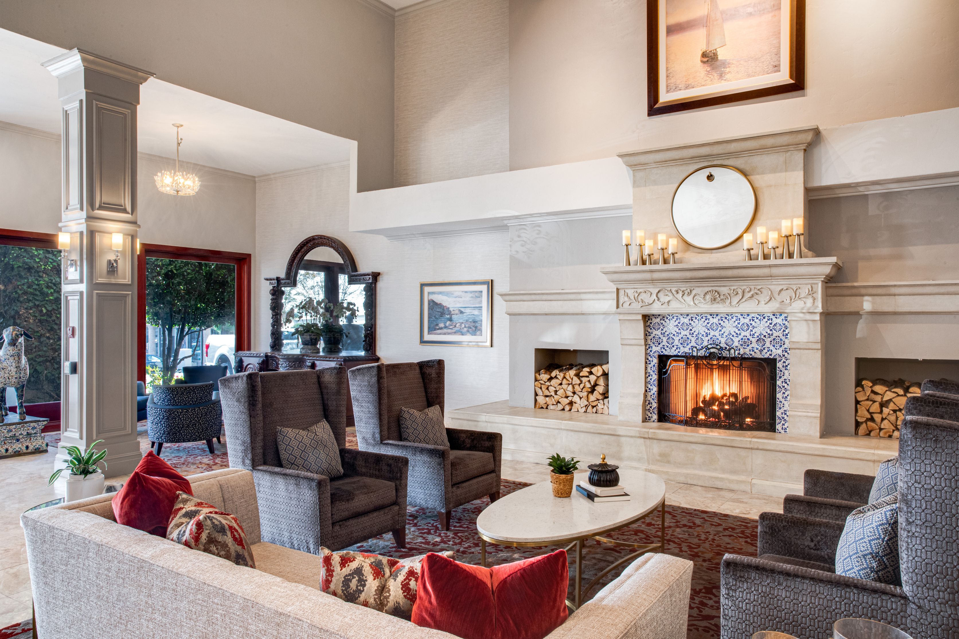 Elegant Ayres Hotels lobby with a lit fireplace, armchairs, sofas with red and beige cushions, a marble coffee table, and decorative plants. Large windows let in natural light; a painting and mirror decorate the walls.