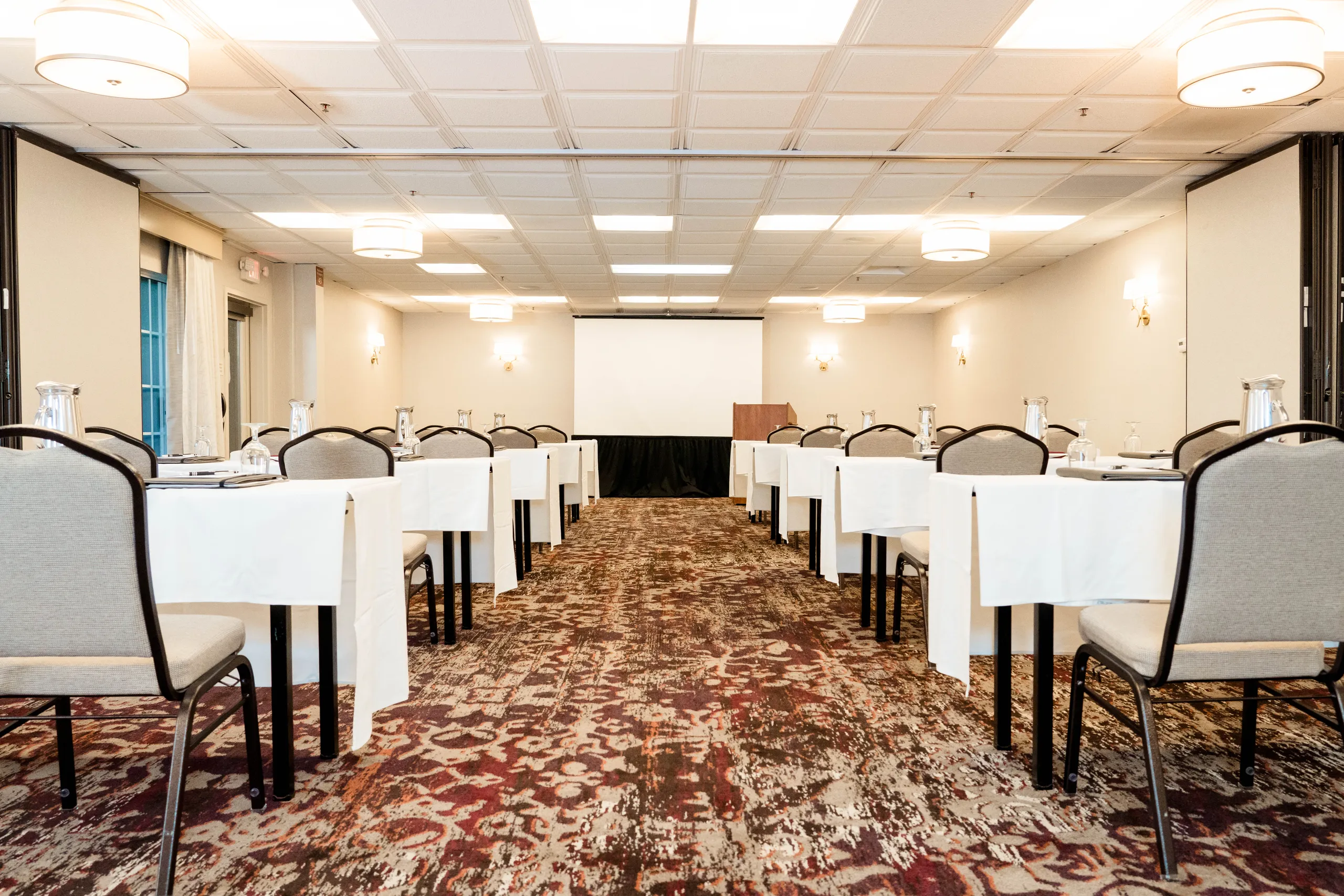 A conference room at Ayres Hotels with rows of tables and chairs facing a podium and projection screen, set up for a meeting or presentation. Water glasses, notepads, and pens are placed on each table.