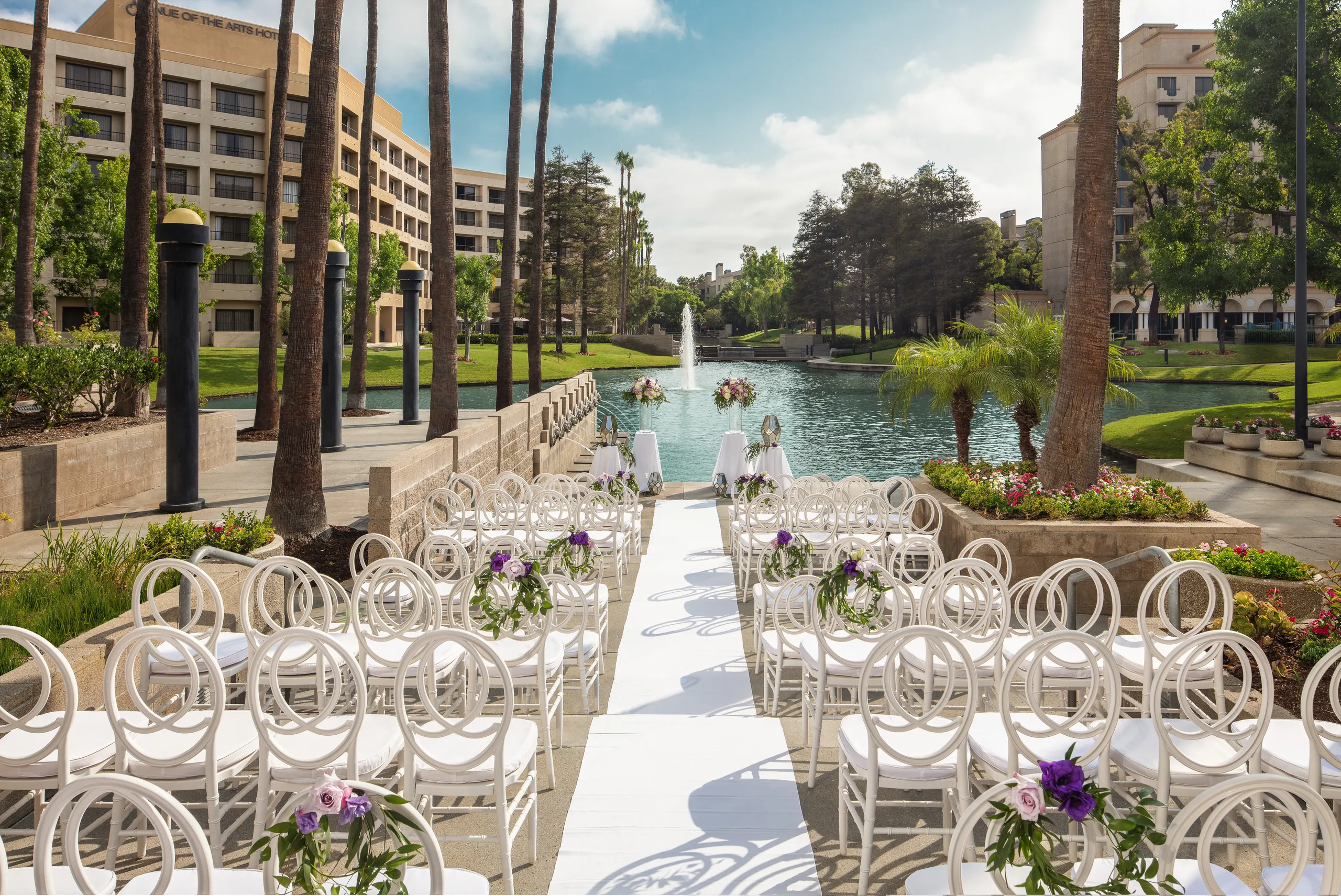 Rows of white chairs with purple flowers face a lake and fountain, set up outdoors at one of the elegant event venues in Costa Mesa for a wedding ceremony between palm trees and buildings under a partly cloudy sky.
