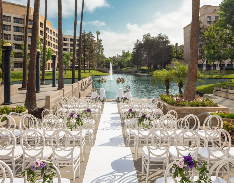 Rows of white chairs with purple flower decorations line an aisle facing a pond and fountain, surrounded by palm trees and buildings under a partly cloudy sky—one of the picturesque wedding venues Costa Mesa has to offer.