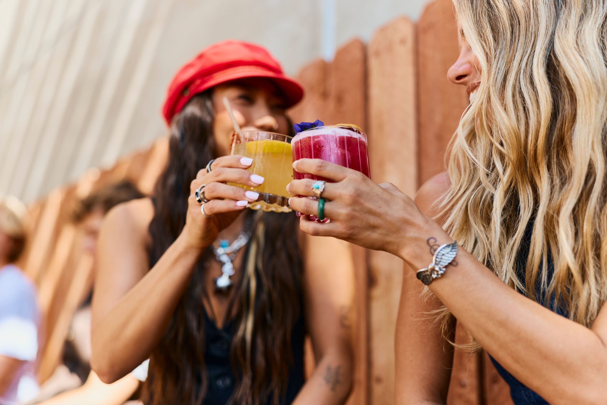 Two women clink colorful cocktails together while smiling outdoors during one of the annual events Costa Mesa is known for. One wears a red cap; the other, with long blonde hair and rings, stands beside a wooden fence in the background.