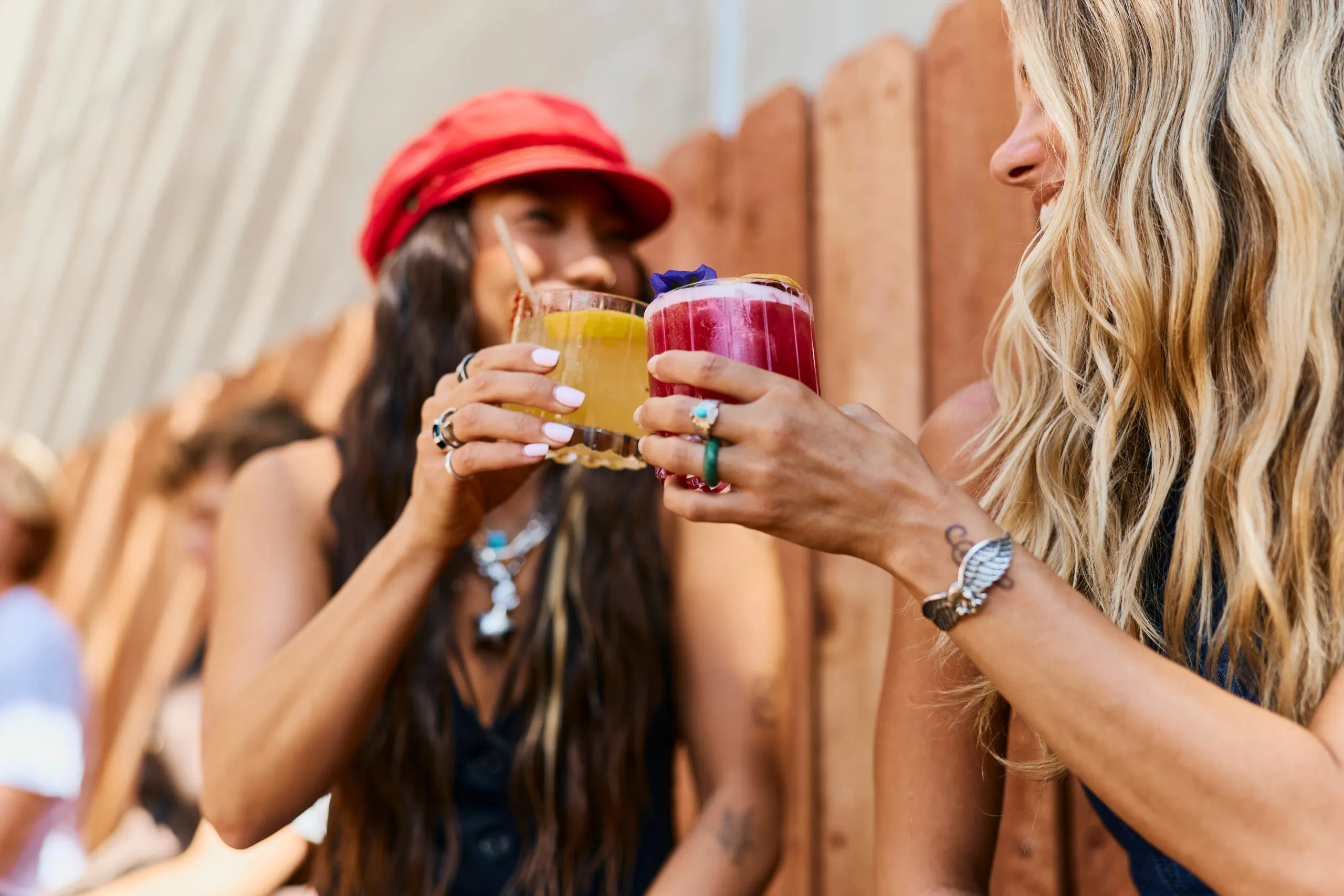 Two women clink colorful cocktails together while smiling outdoors during one of the annual events Costa Mesa is known for. One wears a red cap; the other, with long blonde hair and rings, stands beside a wooden fence in the background.