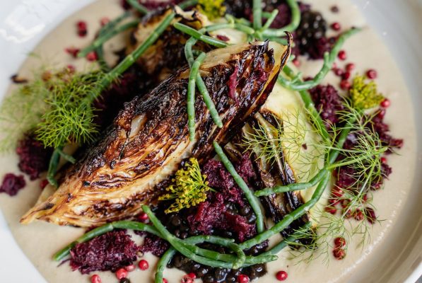 Grilled cabbage wedges on a creamy white sauce, garnished with fresh herbs, green beans, pink peppercorns, and crumbled purple vegetable pieces—an artful dish inspired by the vibrant flavors of Eatcation Costa Mesa.
