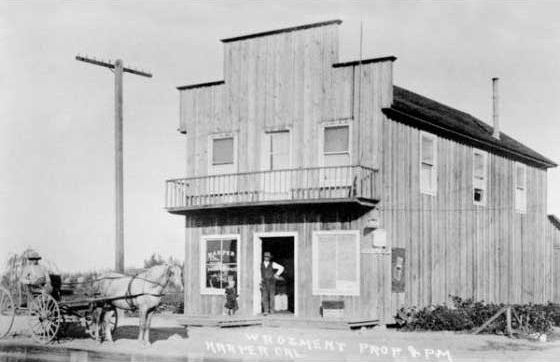 Black and white photo of a two-story wooden building with a balcony, a person in the doorway, and a horse-drawn carriage in front—echoing scenes from the early history of Costa Mesa. The sign reads W. Roement Prop & PM, Hamburg, N.D.