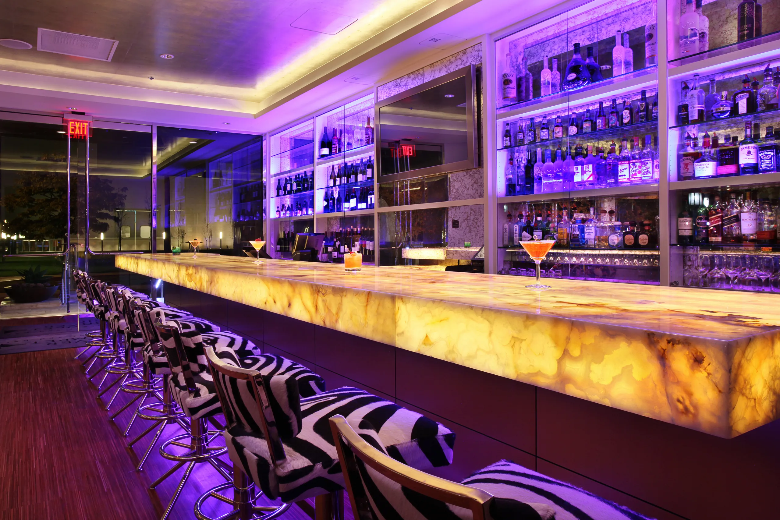 A modern bar at Silver Trumpet Restaurant Costa Mesa features zebra-patterned stools, a glowing onyx counter, and shelves of liquor bottles, all lit with purple lighting. Two cocktails sit on the counter.