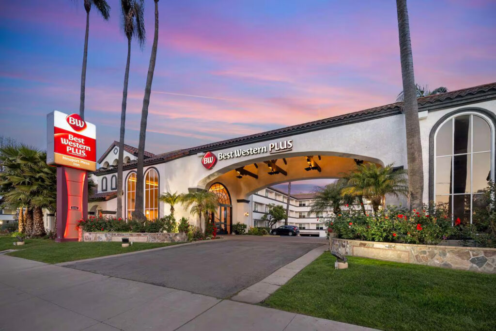 The exterior of the Best Western Costa Mesa Plus hotel at sunset features palm trees, landscaped gardens, arched windows, and a prominent red and white hotel sign welcoming guests at the entrance.